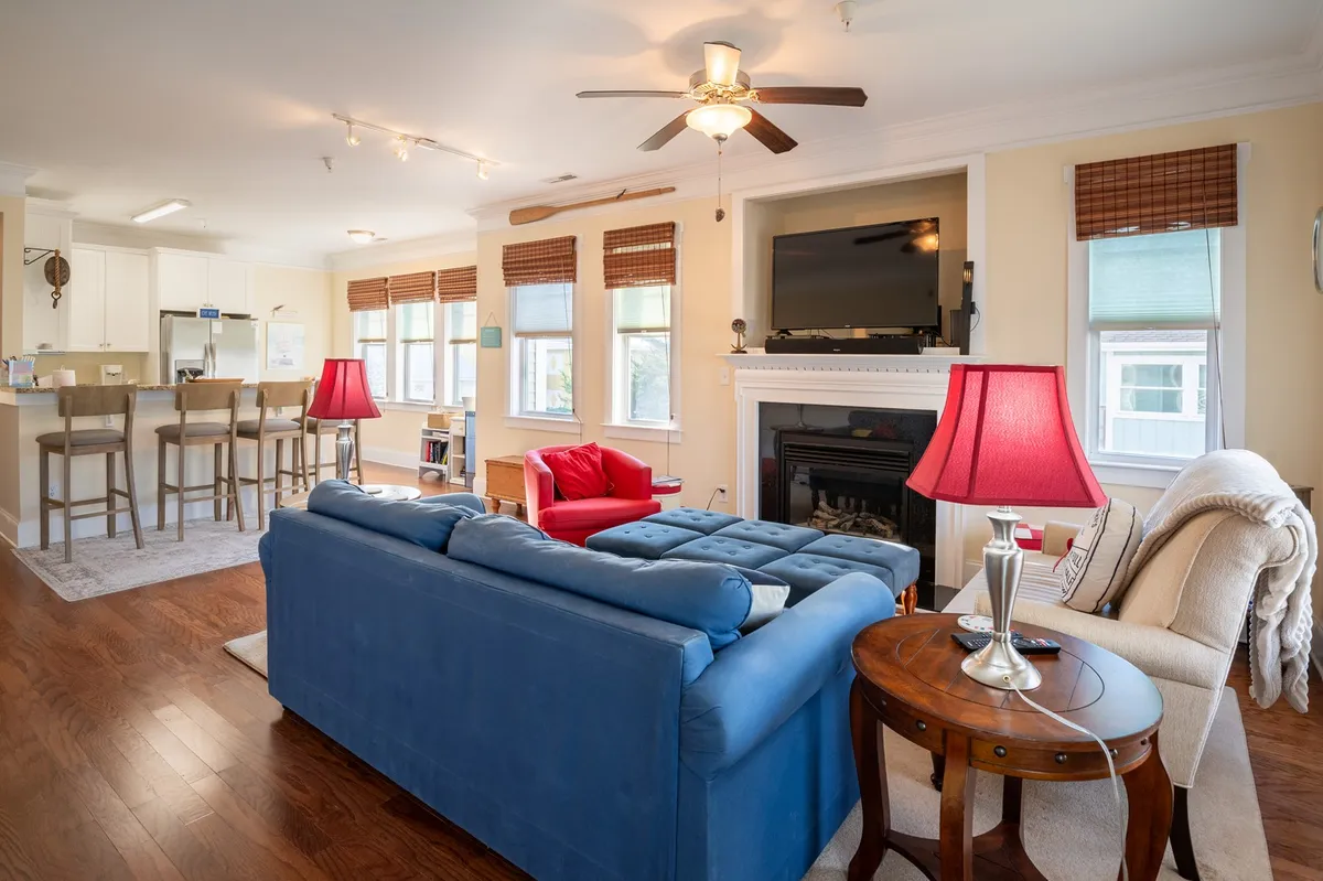 This image captures a cozy living room featuring a blue sofa and red armchair centered around a decorative fireplace. The space also includes bar stools, a wall-mounted television, and ample natural light, evoking a welcoming and comfortable atmosphere.