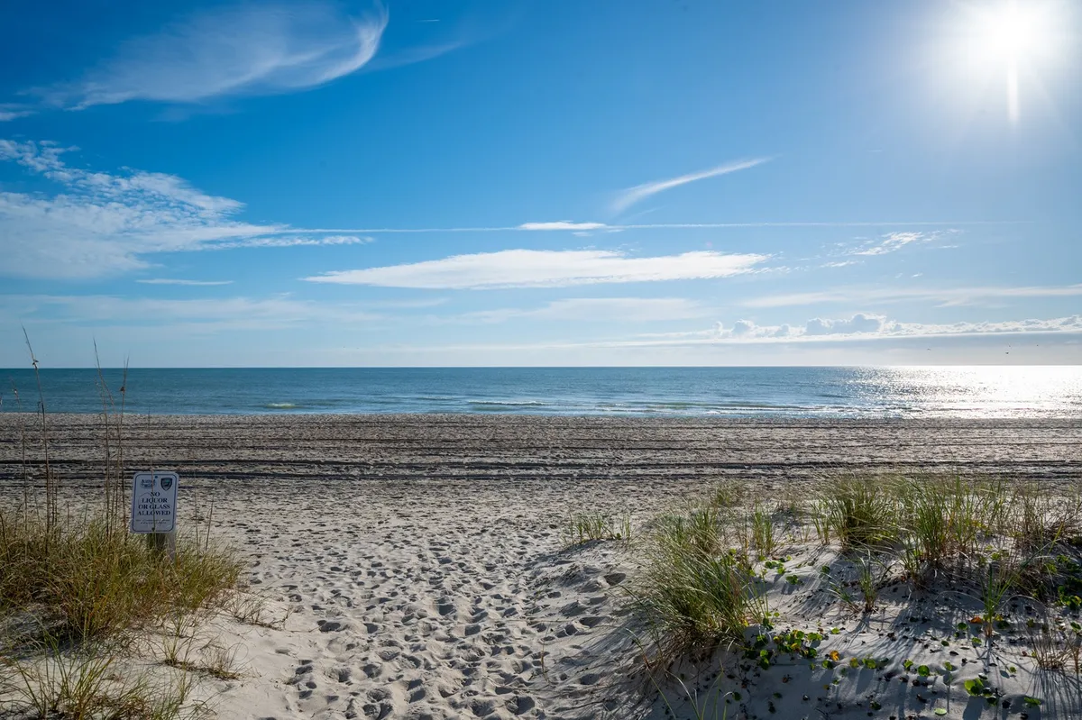 A wide, tranquil stretch of sandy beach bordered by gentle dune grass and the blue ocean under a clear sky. There are no people in the frame, emphasizing the natural beauty and inviting atmosphere of the shoreline.