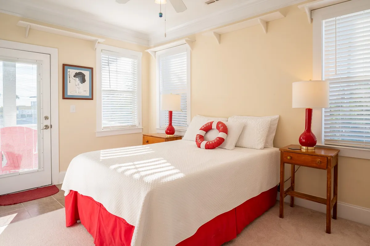 A bedroom featuring a bed with red accents, red lamps, a lifebuoy decoration on the wall, and windows allowing natural light to brighten the space. The bedding is crisp and inviting, with a sense of comfort and tranquility.