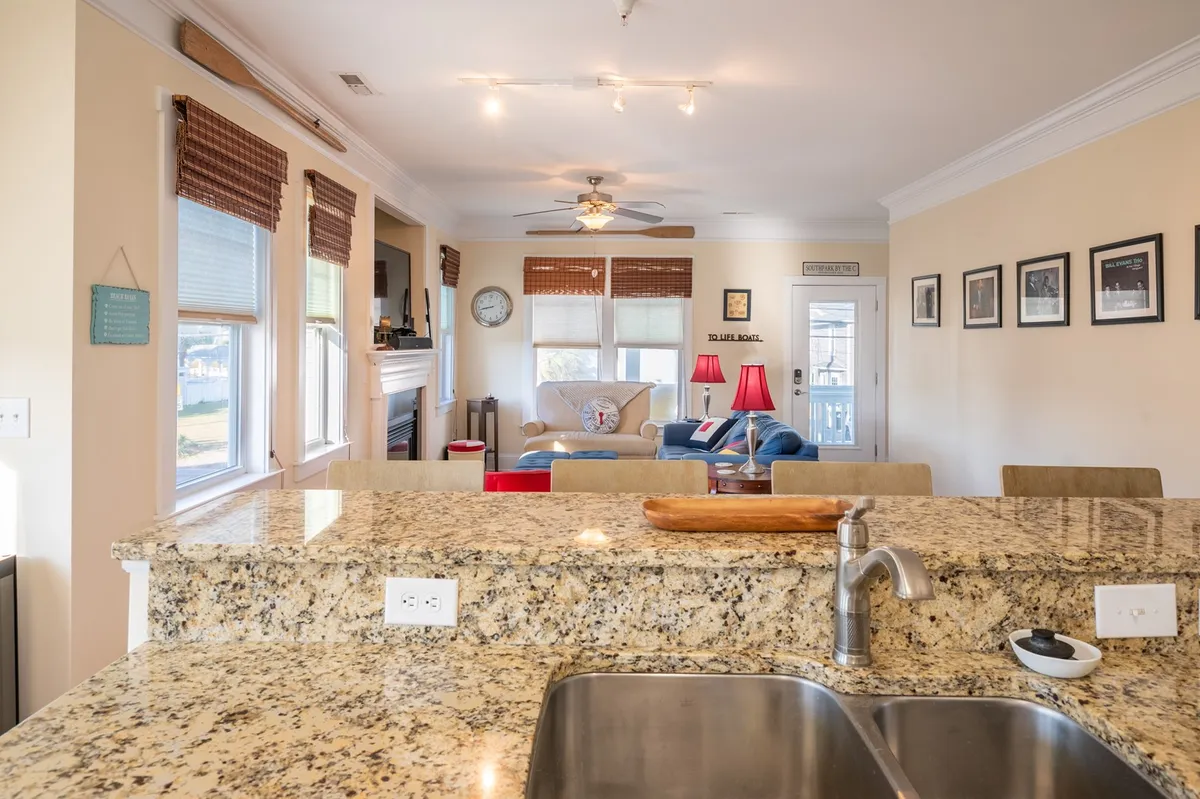 This image presents an open view from the kitchen to the living room, highlighting a granite countertop with a kitchen sink, a comfortable seating area, red lamps, and a wall with family photos. The scene feels warm, with the layout flowing seamlessly from one area to another.