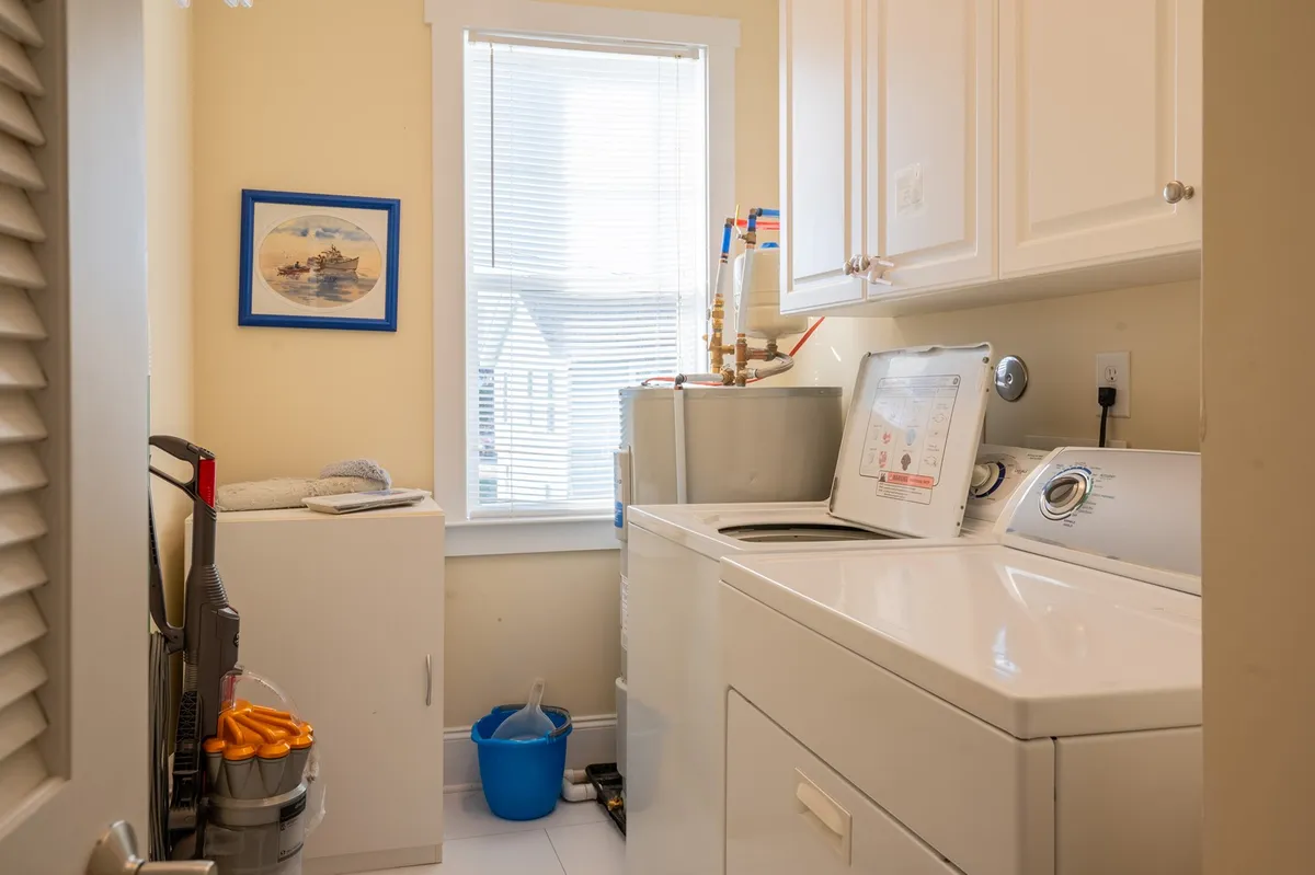 This image features a neatly organized laundry area with a washing machine and dryer, illuminated by natural light from a nearby window. A framed picture on the wall adds a welcoming touch, and the entire space feels clean and functional.