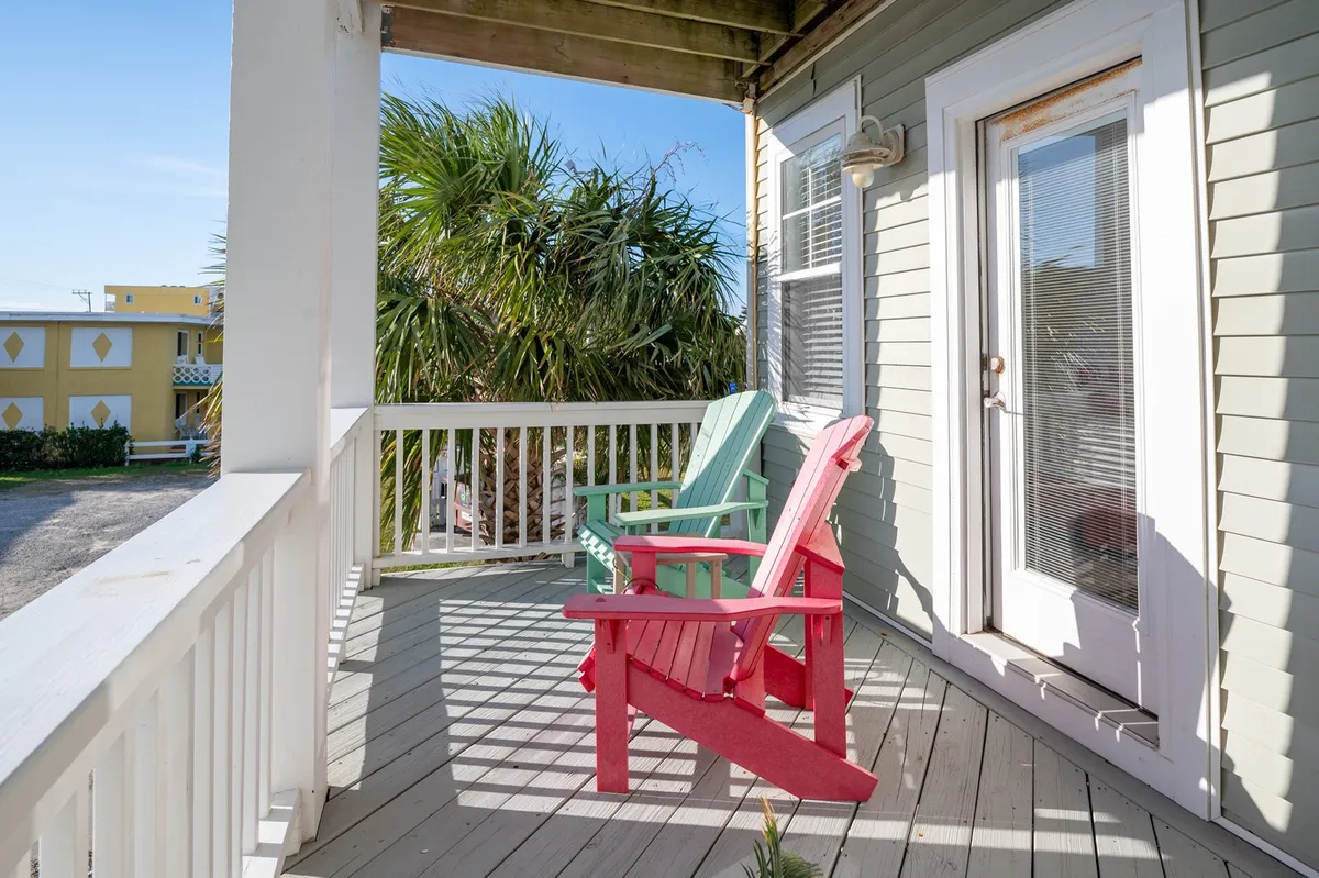 A cheerful exterior view featuring red and green Adirondack chairs set on a private porch surrounded by palm trees, with the doorway to the unit visible. The inviting setup suggests a tranquil spot to relax and enjoy the coastal atmosphere mere steps from the entrance.