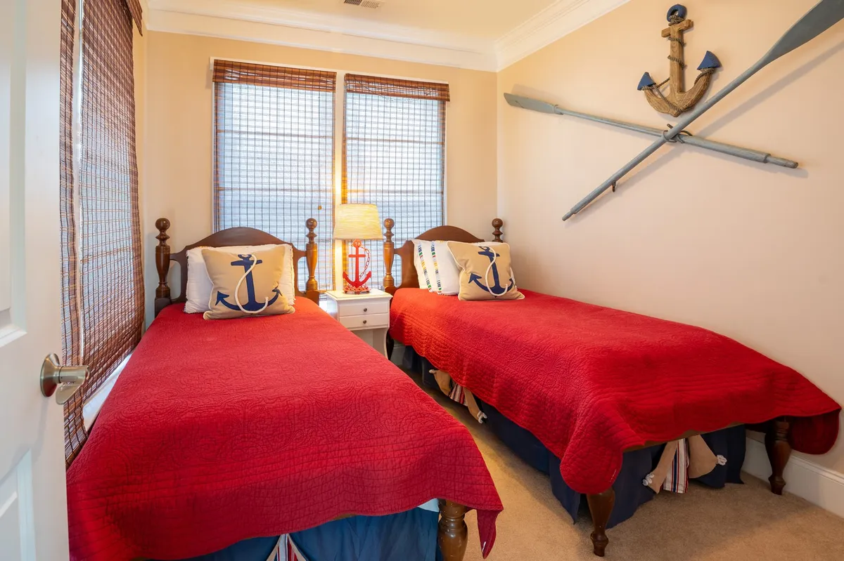 A playful twin bedroom with two twin beds adorned in red quilts and decorative anchor pillows. The room is brightened by a window with blinds and nautical-themed wall décor, creating a cheerful and welcoming space ideal for children or friends.