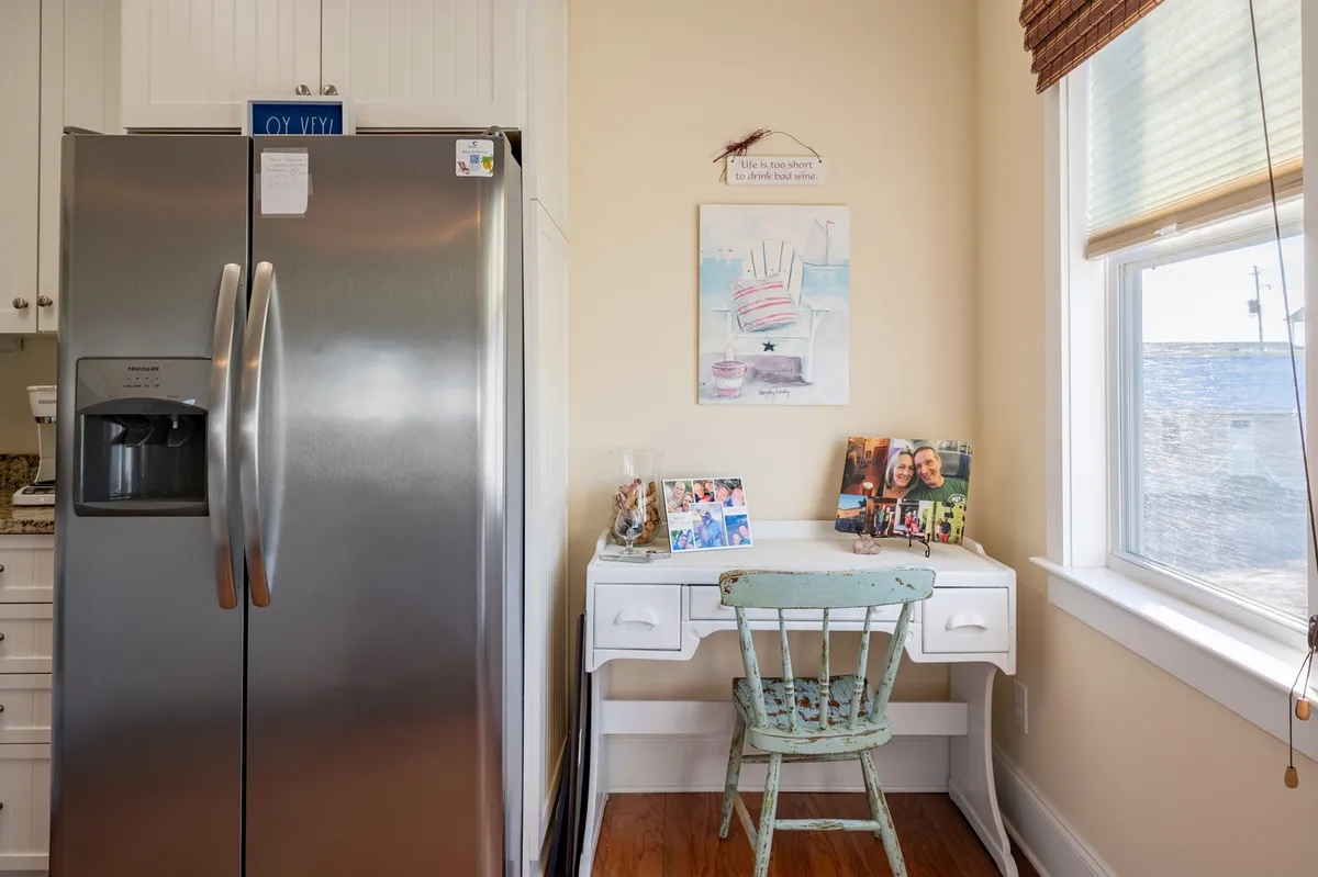 A corner of the home with a stainless steel refrigerator, a white desk featuring a green chair, and family photos and colorful artwork. The window in the background brings in natural light, hinting at a home base filled with warmth and personal touches.